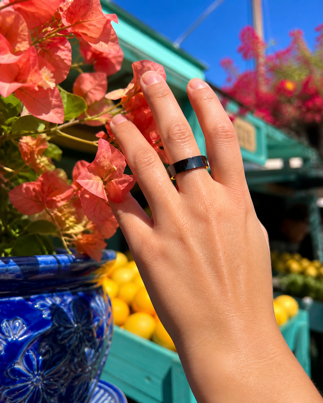 Hand wearing the black Rune 1 ring beside coral flowers and citrus at a colorful market